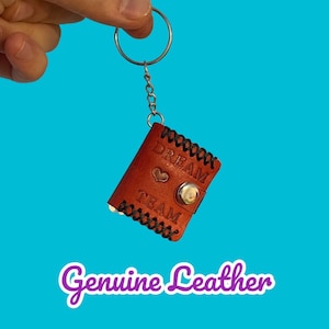 May include: A brown leather keychain with a silver key ring and a small heart charm. The keychain has the words "Dream Team" embossed on it. The text "Genuine Leather" is written in purple below the keychain.