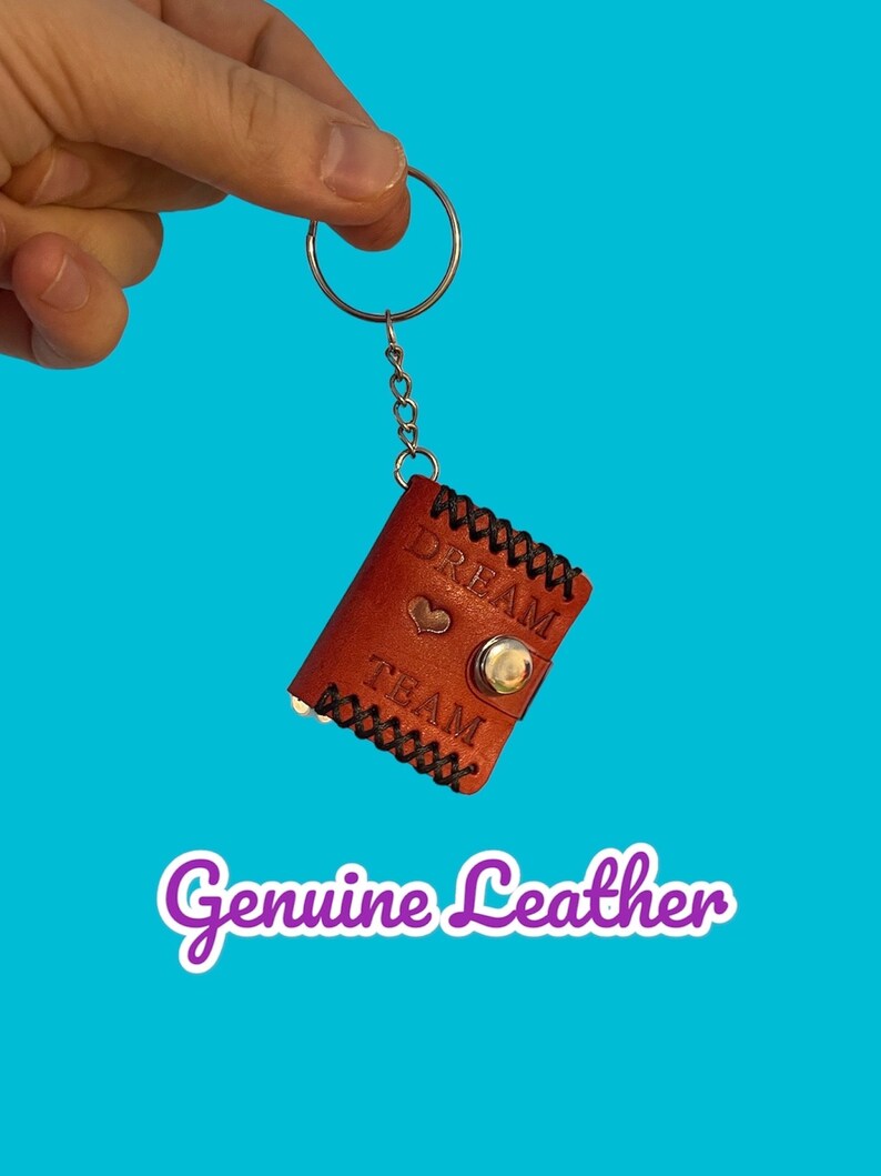 May include: A brown leather keychain with a silver key ring and a small heart charm. The keychain has the words "Dream Team" embossed on it. The text "Genuine Leather" is written in purple below the keychain.