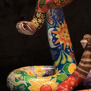 May include: A colorful, hand-painted ceramic snake sculpture with a bright blue, green, yellow, red, and orange floral design. The snake is coiled in a circle with its head raised and its tail curled around its body.