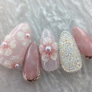 May include: Five pink and white nail tips with different designs. One nail tip has a floral design with a pearl accent, another has a glitter design, and the other three have a pink glitter design.