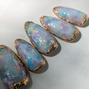 Puede incluir: Cinco dise&ntilde;os de arte de u&ntilde;as ovalados. Cada u&ntilde;a presenta un dise&ntilde;o brillante, iridiscente azul y morado con un borde dorado. Las u&ntilde;as est&aacute;n dispuestas en diagonal sobre un fondo azul claro.