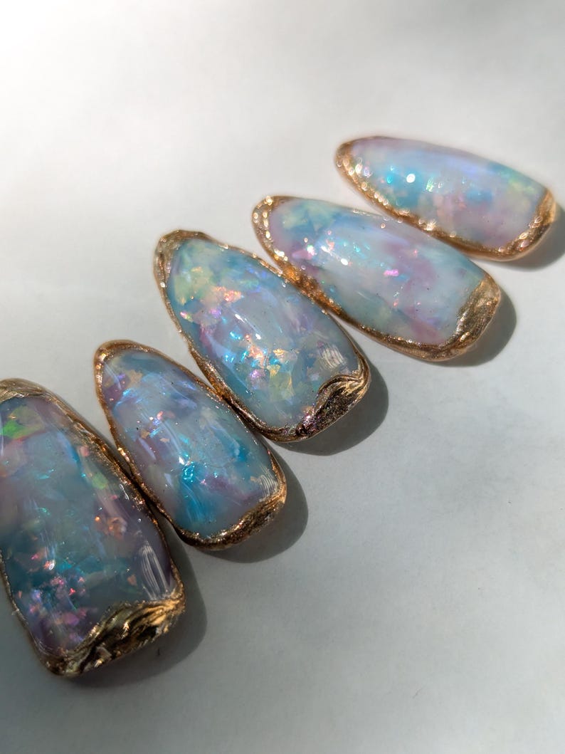 Puede incluir: Cinco dise&ntilde;os de arte de u&ntilde;as ovalados. Cada u&ntilde;a presenta un dise&ntilde;o brillante, iridiscente azul y morado con un borde dorado. Las u&ntilde;as est&aacute;n dispuestas en diagonal sobre un fondo azul claro.