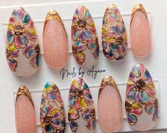 May include: A set of ten artificial nails. Some nails are a light pink with glitter, and others feature a floral design with gold outlines and a variety of colors, including pink, yellow, blue, and purple. The text "Nails by Ayano" is visible.
