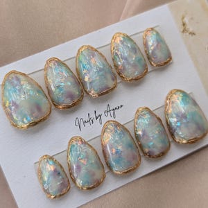 Puede incluir: Un juego de diez u&ntilde;as postizas iridiscentes, de forma ovalada, con un borde met&aacute;lico dorado. Las u&ntilde;as tienen un efecto nacarado, opalescente, con toques de azul, morado y rosa. El texto "Nails by Ayana" est&aacute; escrito en la tarjeta.