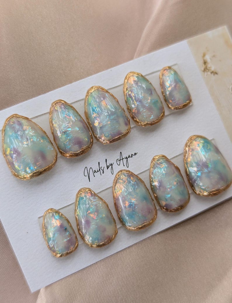 Puede incluir: Un juego de diez u&ntilde;as postizas iridiscentes, de forma ovalada, con un borde met&aacute;lico dorado. Las u&ntilde;as tienen un efecto nacarado, opalescente, con toques de azul, morado y rosa. El texto "Nails by Ayana" est&aacute; escrito en la tarjeta.