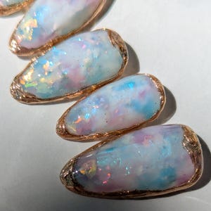 Puede incluir: Primer plano de cinco dise&ntilde;os de u&ntilde;as ovalados e iridiscentes. Cada u&ntilde;a presenta una mezcla de tonos pastel azul, rosa y blanco con brillantes acentos de purpurina. Los bordes est&aacute;n delineados con un borde dorado met&aacute;lico.