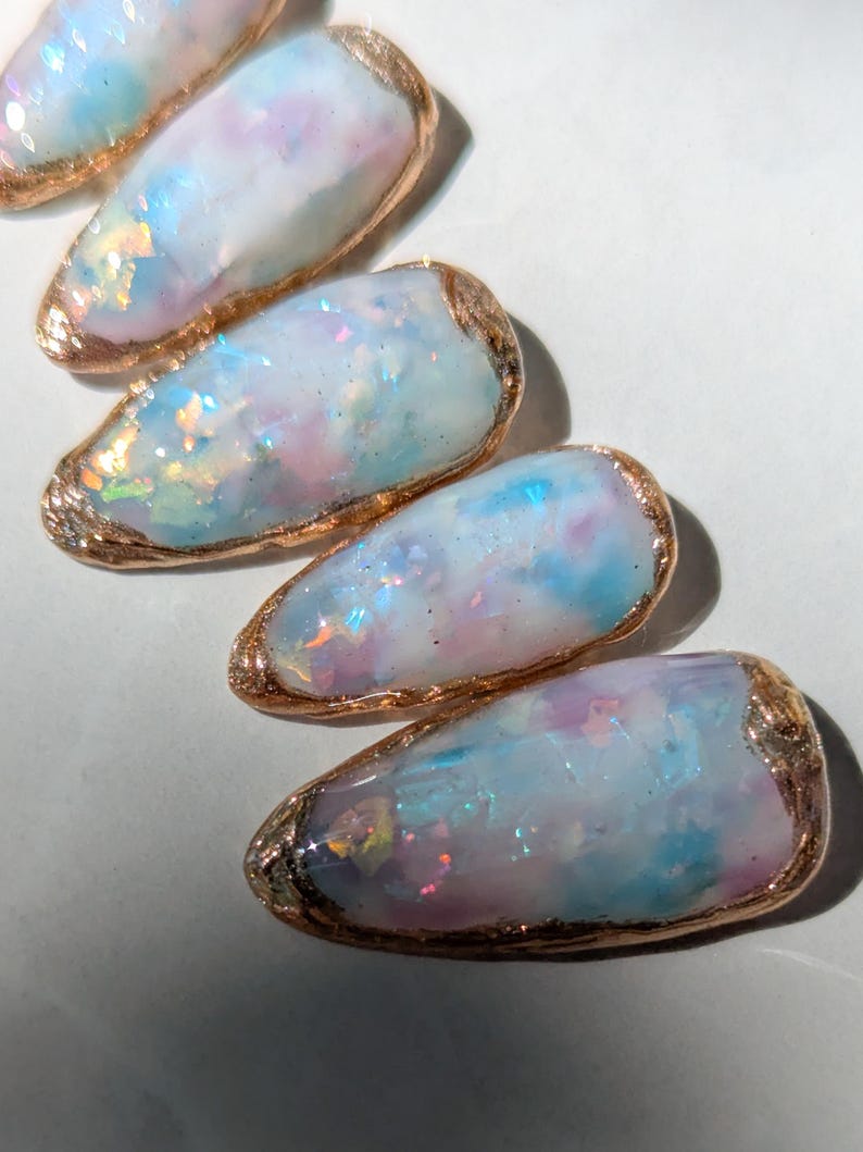 Puede incluir: Primer plano de cinco dise&ntilde;os de u&ntilde;as ovalados e iridiscentes. Cada u&ntilde;a presenta una mezcla de tonos pastel azul, rosa y blanco con brillantes acentos de purpurina. Los bordes est&aacute;n delineados con un borde dorado met&aacute;lico.