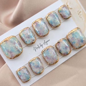 Puede incluir: Un conjunto de diez u&ntilde;as postizas cuadradas. Cada u&ntilde;a presenta un dise&ntilde;o iridiscente y brillante en tonos azules, morados y blancos, enmarcado con un borde met&aacute;lico dorado. El texto "Nails by Ayana" es visible en la tarjeta.