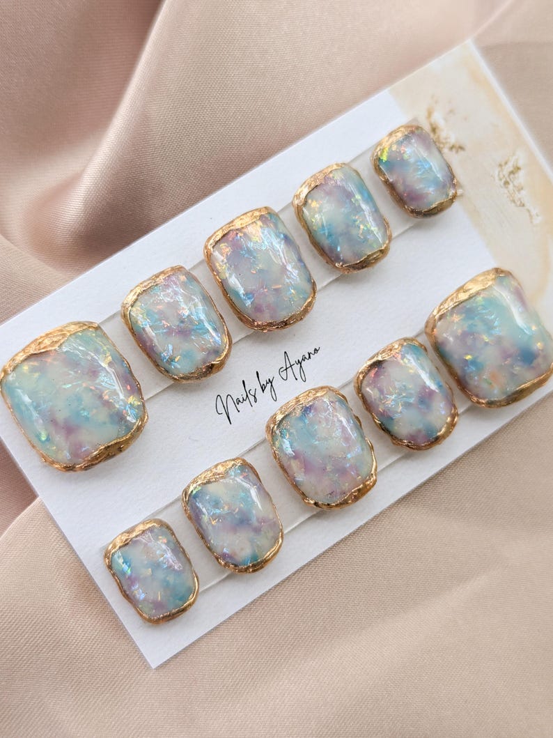 Puede incluir: Un conjunto de diez u&ntilde;as postizas cuadradas. Cada u&ntilde;a presenta un dise&ntilde;o iridiscente y brillante en tonos azules, morados y blancos, enmarcado con un borde met&aacute;lico dorado. El texto "Nails by Ayana" es visible en la tarjeta.