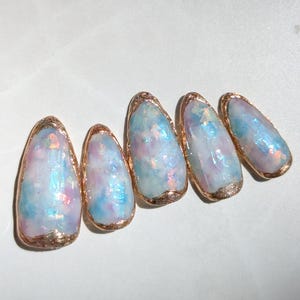 Puede incluir: Cinco u&ntilde;as artificiales de forma ovalada con un dise&ntilde;o iridiscente. Las u&ntilde;as presentan una mezcla de tonos azules, rosas y blancos, con un borde dorado en los bordes. Las u&ntilde;as est&aacute;n dispuestas en fila, mostrando su apariencia art&iacute;stica &uacute;nica.