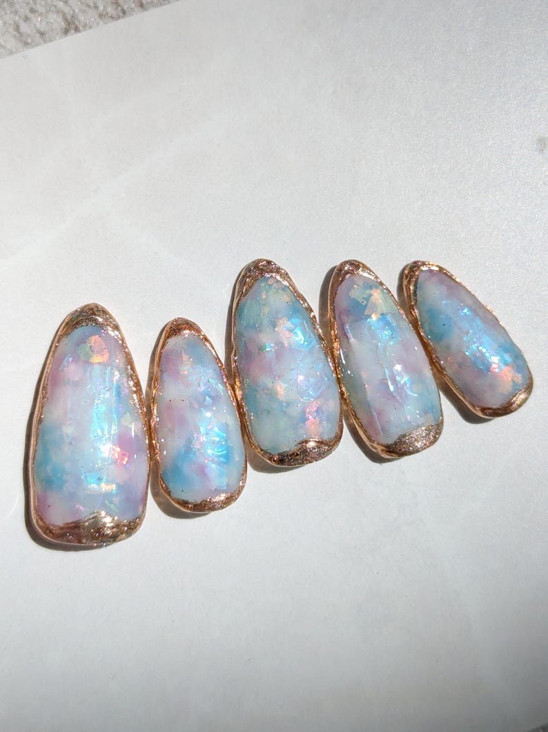 Puede incluir: Cinco u&ntilde;as artificiales de forma ovalada con un dise&ntilde;o iridiscente. Las u&ntilde;as presentan una mezcla de tonos azules, rosas y blancos, con un borde dorado en los bordes. Las u&ntilde;as est&aacute;n dispuestas en fila, mostrando su apariencia art&iacute;stica &uacute;nica.