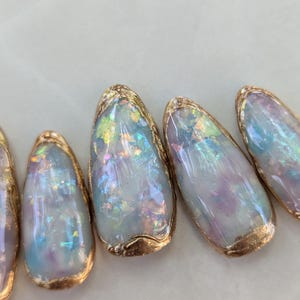 Puede incluir: Primer plano de varias u&ntilde;as ovaladas iridiscentes con un borde met&aacute;lico dorado. Las u&ntilde;as tienen un efecto nacarado y opalino con tonos de azul, morado y verde. El borde dorado a&ntilde;ade un toque de elegancia al dise&ntilde;o.