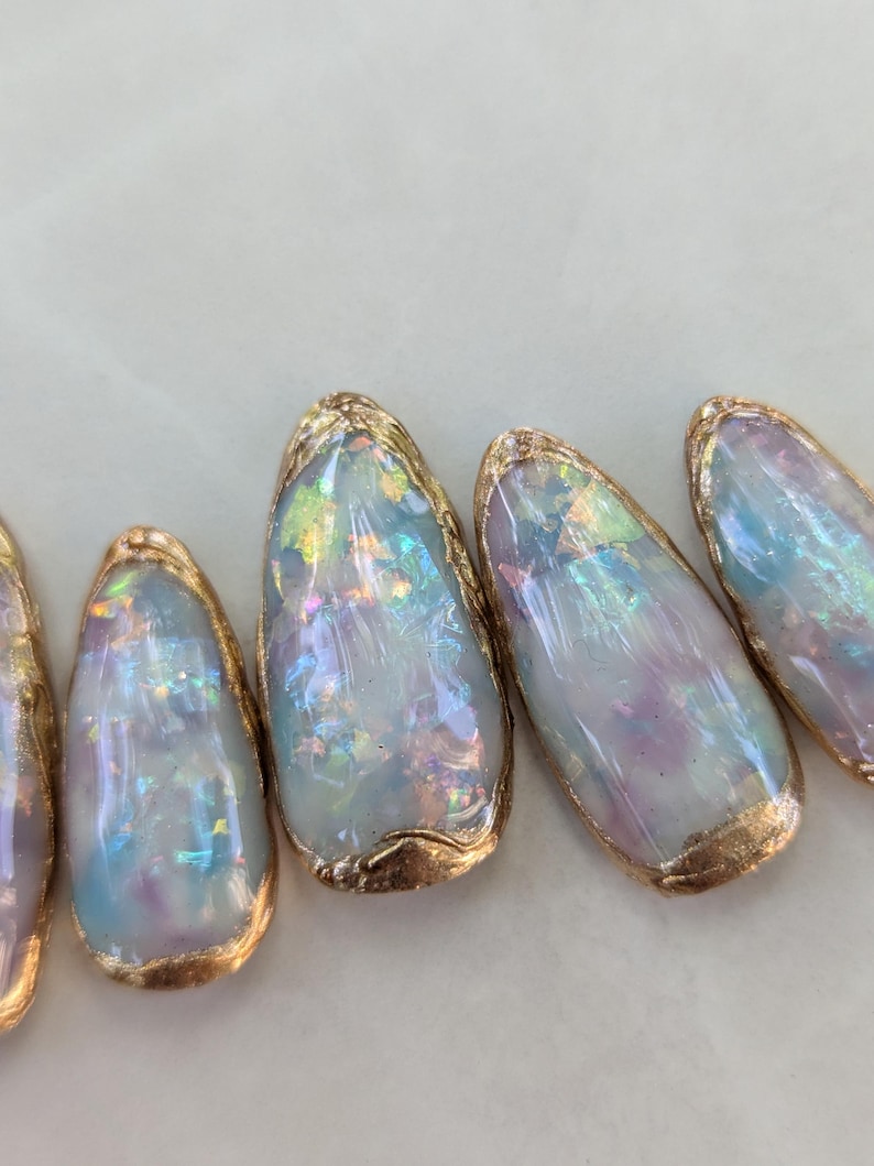 Puede incluir: Primer plano de varias u&ntilde;as ovaladas iridiscentes con un borde met&aacute;lico dorado. Las u&ntilde;as tienen un efecto nacarado y opalino con tonos de azul, morado y verde. El borde dorado a&ntilde;ade un toque de elegancia al dise&ntilde;o.