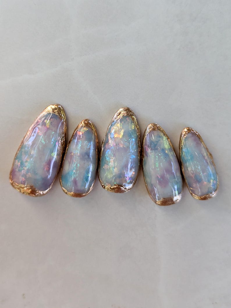 Puede incluir: Cinco puntas de u&ntilde;as ovaladas iridiscentes con un borde met&aacute;lico dorado. Las u&ntilde;as tienen un efecto nacarado y opalescente con tonos de azul, morado y rosa. El borde dorado a&ntilde;ade un toque de elegancia al arte de u&ntilde;as.