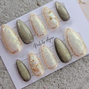 May include: Set of ten press-on nails with a gold glitter and iridescent white finish. Some nails have gold accents and small gold and white embellishments. The nails are displayed on a white card with the text "Nails by Ayana".