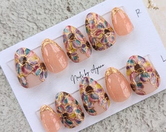 May include: A set of ten press-on nails arranged on a white display card labeled with R and L. The nails feature a peach-colored base with fine glitter, accented by floral patterns in shades of purple, green, and yellow outlined in gold foil.