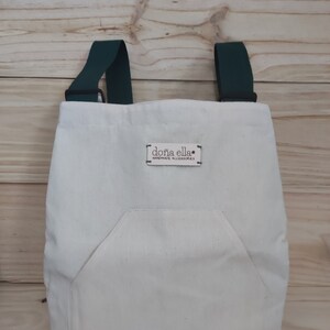 May include: A white canvas tote bag with a green strap and a pocket on the front. The bag has a brown leather label that says "doña ella* handmade accessories".