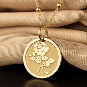 May include: A gold necklace with a round pendant featuring an engraved rose design. The pendant is on a chain with small round beads.