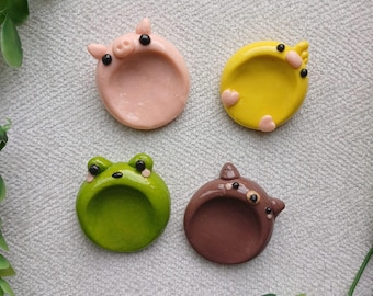 May include: Four handmade ceramic ring dishes in the shape of animals: a pink pig, a yellow chick, a green frog, and a brown bear. Each dish has a small, raised animal head and a circular indentation for holding rings or small items.