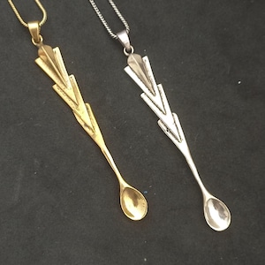 May include: Two delicate necklaces with spoon pendants. One necklace has a gold-toned pendant and the other has a silver-toned pendant. Both pendants feature a unique, geometric design.