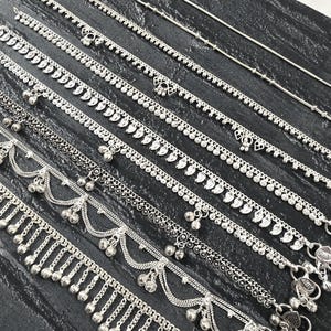 May include: A collection of silver-toned anklets arranged in parallel, featuring various designs including delicate chains, intricate leaf-shaped charms, layered strands with hanging bells, and ornate metalwork with small spherical dangles.
