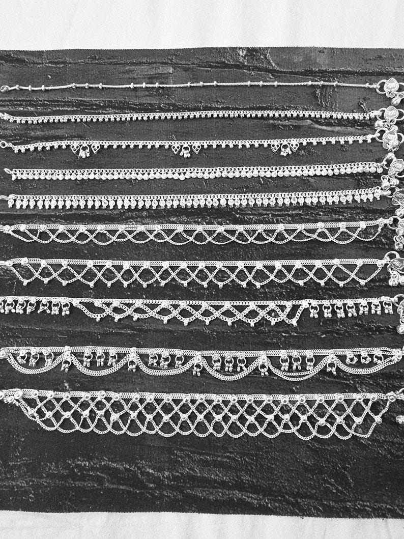 Pode incluir: Uma cole&ccedil;&atilde;o de pulseiras de tornozelo prateadas com v&aacute;rios designs, incluindo correntes de contas, padr&otilde;es delicados e pingentes pendurados. As pulseiras s&atilde;o exibidas em uma superf&iacute;cie escura e texturizada.