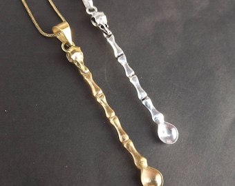 May include: Two silver spoon necklaces, one gold and one silver, hanging against a dark background. The spoons are long and slender with a rounded end.