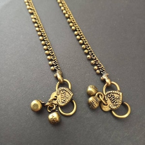 Op de afbeelding: Twee gouden enkelbandjes met hartvormige bedels waarop "Shiv" staat. Elk enkelbandje heeft een kleine belbedel.