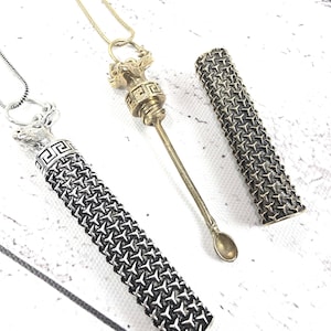 Può includere: Tre collane in metallo con ciondoli decorativi e custodie cilindriche abbinate. Una collana presenta un ciondolo a forma di cucchiaio, mentre le altre hanno disegni geometrici. Le custodie hanno un motivo strutturato.