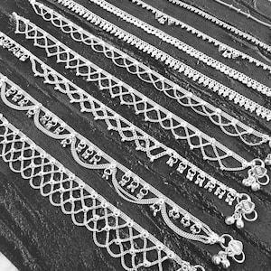 Pode incluir: Uma cole&ccedil;&atilde;o de pulseiras de tornozelo prateadas com v&aacute;rios designs decorativos, incluindo padr&otilde;es de contas, correntes e vazados. Algumas pulseiras de tornozelo apresentam pequenos pingentes pendurados e sinos. As pulseiras s&atilde;o exibidas em uma superf&iacute;cie escura e texturizada.