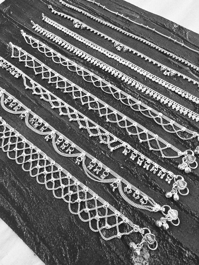 Pode incluir: Uma cole&ccedil;&atilde;o de pulseiras de tornozelo prateadas com v&aacute;rios designs decorativos, incluindo padr&otilde;es de contas, correntes e vazados. Algumas pulseiras de tornozelo apresentam pequenos pingentes pendurados e sinos. As pulseiras s&atilde;o exibidas em uma superf&iacute;cie escura e texturizada.