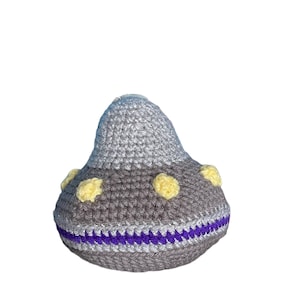 May include: A handmade crochet toy in the shape of a UFO. The top is light gray, the middle is dark gray, and the bottom has a purple stripe. Yellow circles are evenly spaced around the middle section.