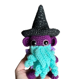 May include: A purple crocheted witch with a black sparkly pointed hat and a turquoise crocheted scarf. The witch has large black eyes with pink eyelids.