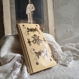 Pu&ograve; includere: Un libro antico color avorio con dettagli floreali e monogrammi in oro e marrone. Il libro &egrave; leggermente aperto. Sullo sfondo una fotografia d'epoca di un ragazzo in divisa da marinaio.