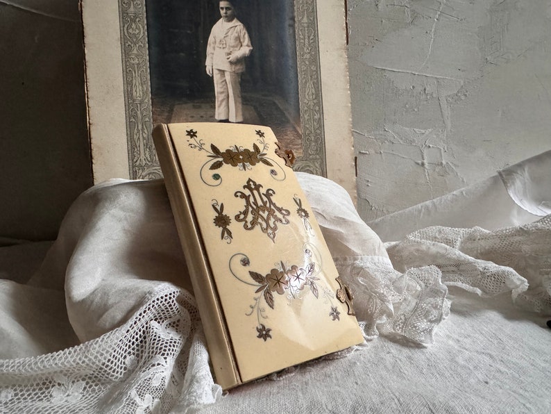 Pu&ograve; includere: Un libro antico color avorio con dettagli floreali e monogrammi in oro e marrone. Il libro &egrave; leggermente aperto. Sullo sfondo una fotografia d'epoca di un ragazzo in divisa da marinaio.