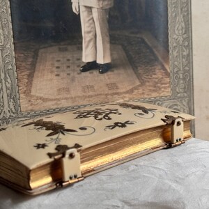 Pu&ograve; includere: Un libro vintage color avorio con bordi dorati e motivi floreali marrone scuro. Il libro &egrave; assicurato con piccole chiusure color crema. Lo sfondo presenta una fotografia in bianco e nero incorniciata.