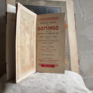 Pu&ograve; includere: Un libro antico aperto, intitolato "PEQUE&Ntilde;O OFICIO DEL DOMINGO" in spagnolo. La copertina &egrave; di un rosso sbiadito con testo nero. Il libro &egrave; aperto su una pagina con testo e un bordo decorativo. Lo sfondo &egrave; una superficie chiara e strutturata.