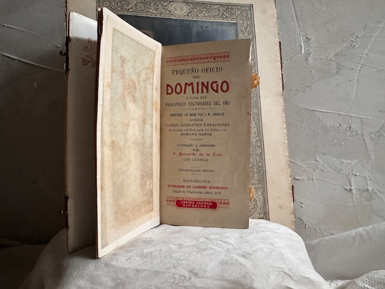 Pu&ograve; includere: Un libro antico aperto, intitolato "PEQUE&Ntilde;O OFICIO DEL DOMINGO" in spagnolo. La copertina &egrave; di un rosso sbiadito con testo nero. Il libro &egrave; aperto su una pagina con testo e un bordo decorativo. Lo sfondo &egrave; una superficie chiara e strutturata.