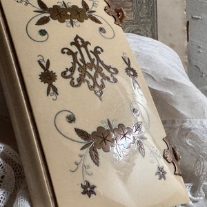 Pu&ograve; includere: Libro antico color crema con decorazioni floreali e monogrammi dorati e argentati. Il libro ha un dorso e angoli dorati. La copertina presenta una finitura lucida.