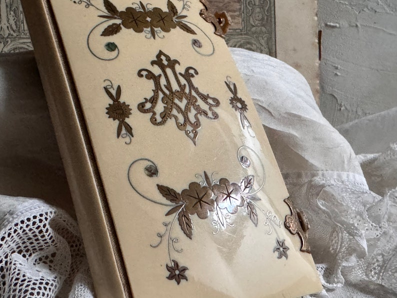 Pu&ograve; includere: Libro antico color crema con decorazioni floreali e monogrammi dorati e argentati. Il libro ha un dorso e angoli dorati. La copertina presenta una finitura lucida.