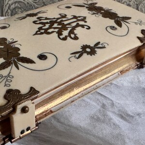 Pu&ograve; includere: Scatola antica color avorio con finiture dorate e decorazioni floreali e a volute. La scatola presenta angoli ornati e una chiusura. Il design include motivi floreali e a volute color oro sul coperchio.