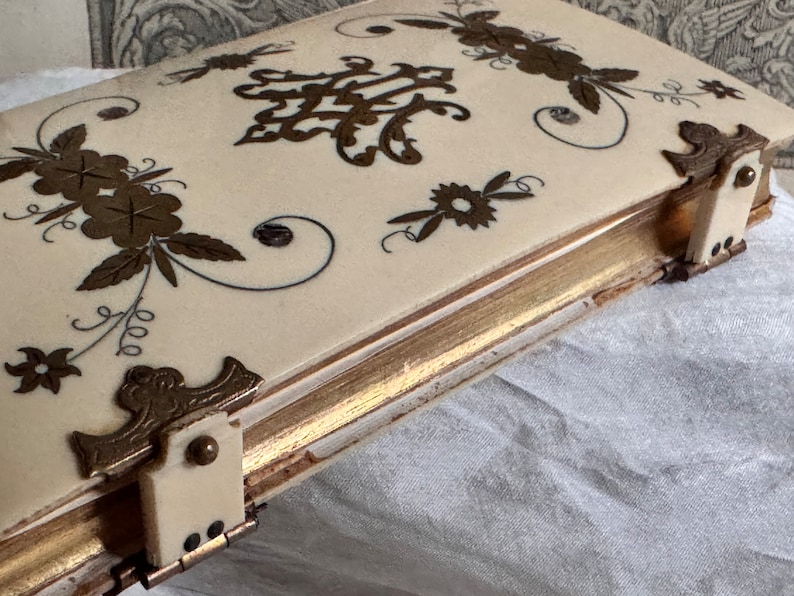 Pu&ograve; includere: Scatola antica color avorio con finiture dorate e decorazioni floreali e a volute. La scatola presenta angoli ornati e una chiusura. Il design include motivi floreali e a volute color oro sul coperchio.