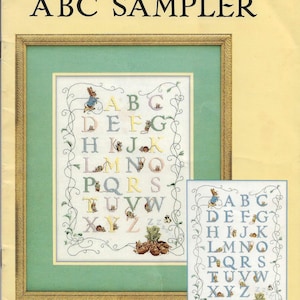 Könnte beinhalten: Ein Beatrix Potter ABC Sampler Kreuzstich-Musterheft mit einem gerahmten Alphabet-Design und kleinen Illustrationen von Kaninchen und Gemüse. Das Cover zeigt den Titel, das Green Apple Co. Logo und den Text Design Adapted by Jeanne Bowers.