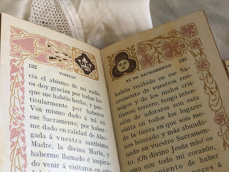 Pu&ograve; includere: Pagine di un libro aperto con testo e bordi decorativi. Le pagine sono numerate 132 e 133. I bordi presentano disegni floreali e di corona in rosa e oro. Il testo &egrave; in spagnolo.