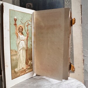 Pu&ograve; includere: Un libro antico aperto con un'illustrazione religiosa sulla pagina sinistra. L'illustrazione raffigura una figura in abiti bianchi che tiene un calice e un bastone. Il libro ha una chiusura dorata e pagine invecchiate.