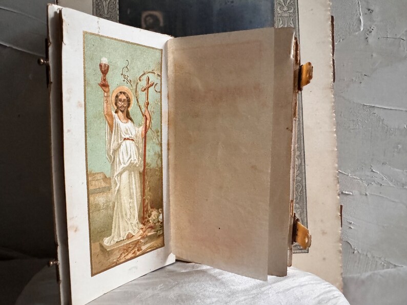 Pu&ograve; includere: Un libro antico aperto con un'illustrazione religiosa sulla pagina sinistra. L'illustrazione raffigura una figura in abiti bianchi che tiene un calice e un bastone. Il libro ha una chiusura dorata e pagine invecchiate.