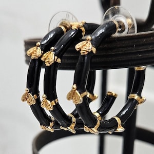 May include: Black hoop earrings with gold bee accents. The earrings are made of black enamel and have a gold-tone finish.