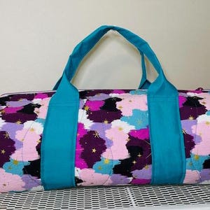 May include: A quilted duffel bag with a vibrant abstract pattern in shades of purple, pink, blue, and gold. The bag features teal handles and accents, offering a stylish and functional design for travel or everyday use.