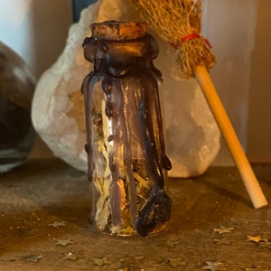 May include: A small glass bottle with a cork stopper, sealed with dark purple wax. The bottle contains dried herbs and flowers. A miniature broom and a white crystal are in the background. Decorative stars are scattered on the surface.