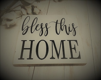 Può includere: Un cartello di legno bianco con lettere nere che recita "bless this home".